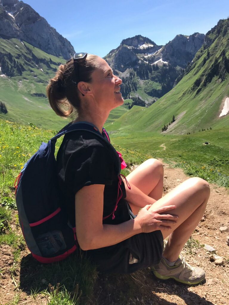 Photographie de Caroline Delhonte Naturopathe, assise sur un rocher au coeur de montagnes verdoyantes. Elle porte un sac à dos, un t-shirt, un shirt et des lunettes sur la tête.